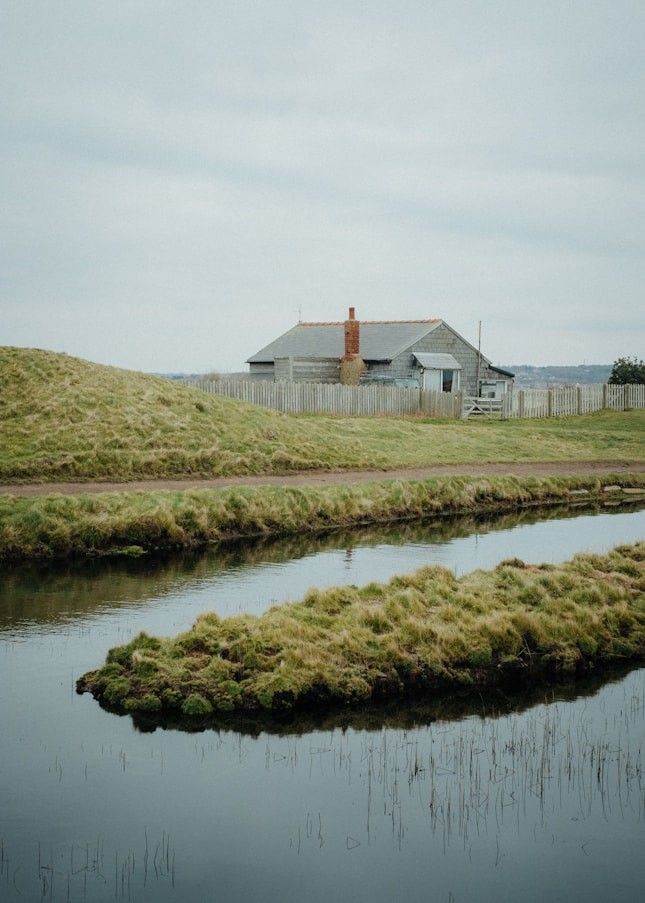 Small weatherboard cottage beyond a grassy river meadow and a curving stream