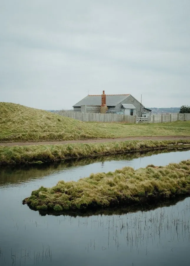 Small weatherboard cottage beyond a grassy river meadow and a curving stream